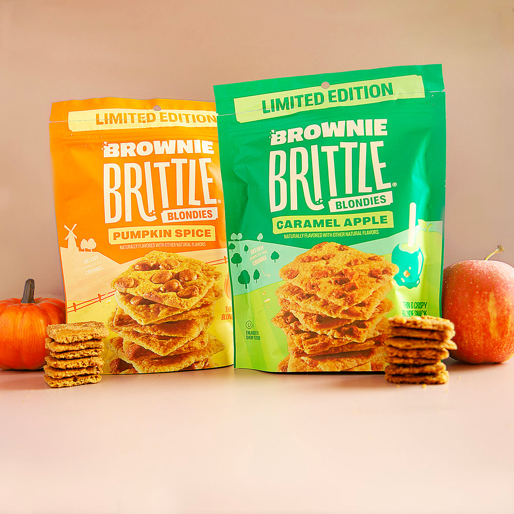 Two packages of Brownie Brittle blondies with pumpkins and an apple on a beige background