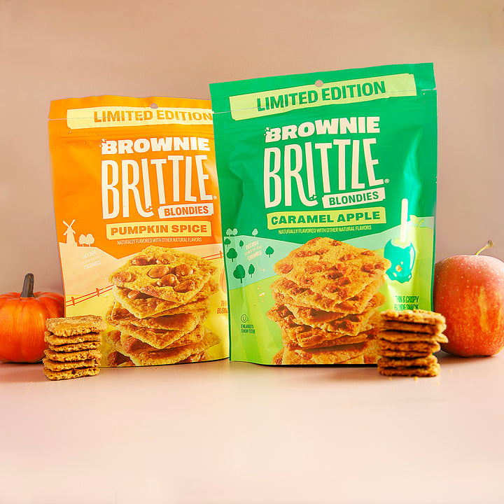 Two packages of Brownie Brittle blondies with pumpkins and an apple on a beige background