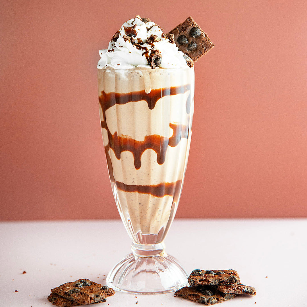 Chocolate milkshake with whipped cream, chocolate drizzle, and cookies on a pink background with brownie brittle on top