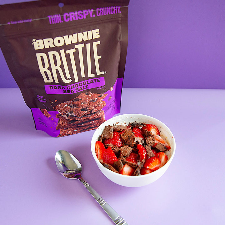 Brownie Brittle packaging with a bowl of brownie pieces and strawberries on a purple background