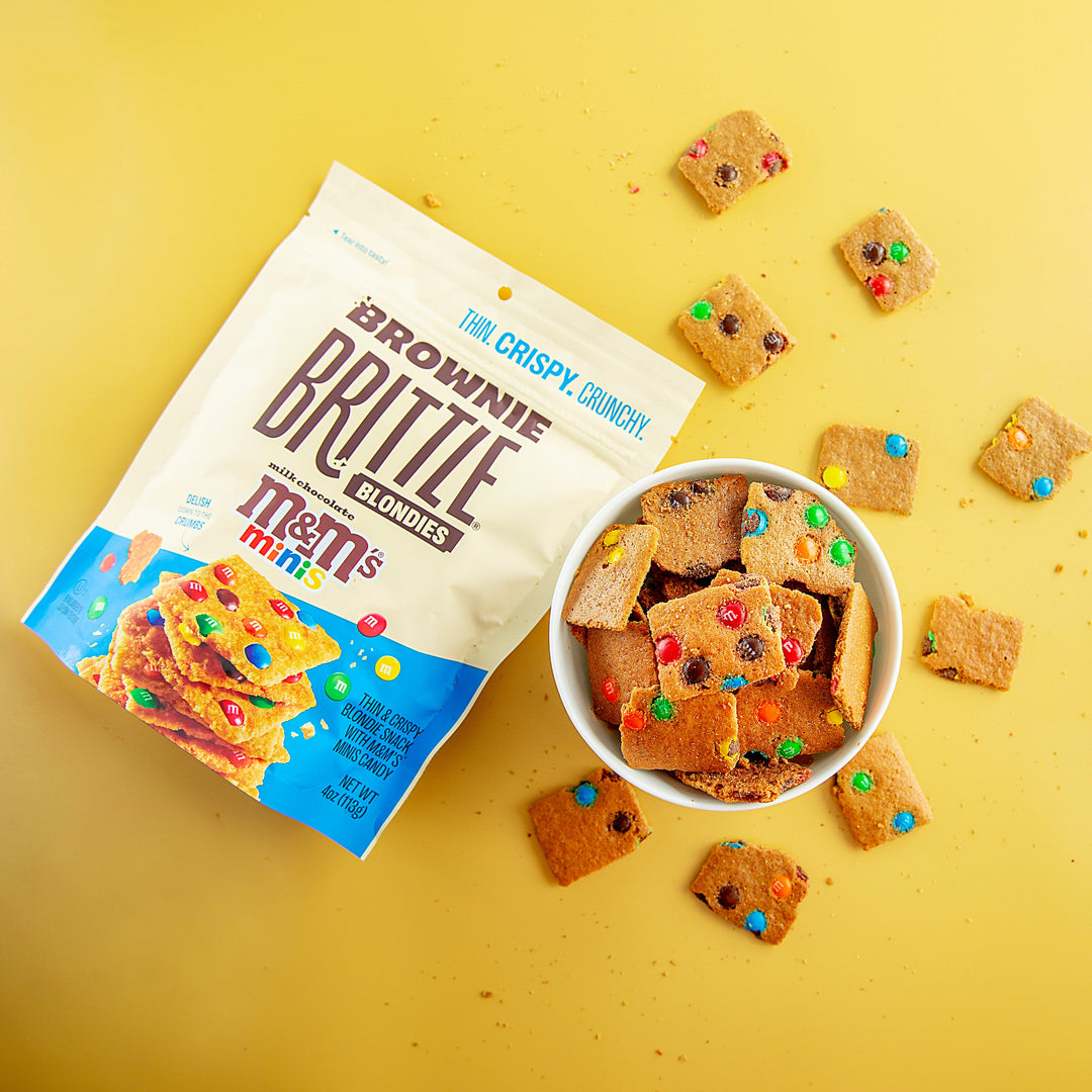a yellow table with M&M'S® Minis Blondie Brownie Brittle in a bowl next to the package