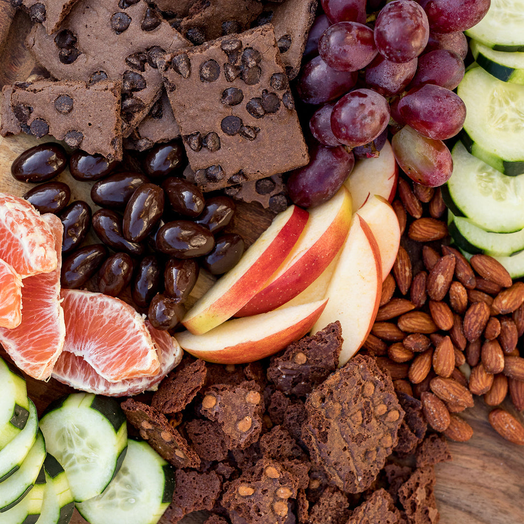 Assorted snack platter with brownie brittle, fruits, and nuts on a wooden board.