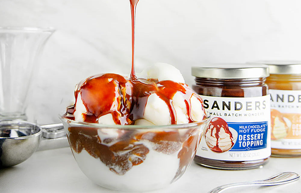 Bowl of ice cream with Sanders hot fudge being poured over it, next to a jar of Sanders Hot Fudge Dessert Topping.