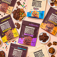 Assorted Brownie Brittle packages are arranged on a light pink background. Bowls of candies and brownie pieces are scattered, creating a playful, indulgent vibe.