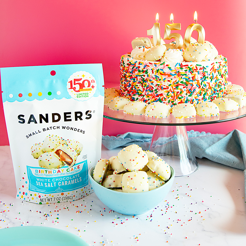  Sanders birthday cake caramels with small batch wonders packaging and a birthday cake in the background.