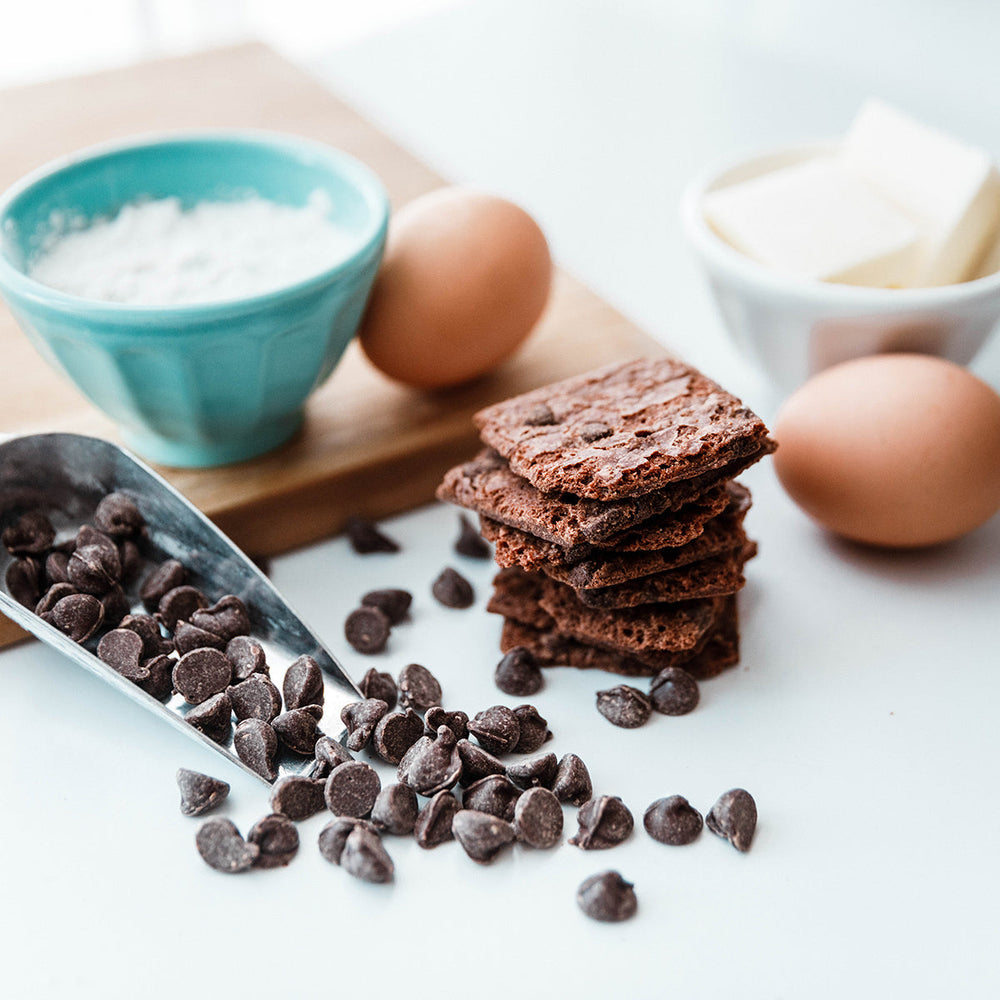 Chocolate chips, stacked brownie brittle, eggs, and a bowl of flour on a white surface.