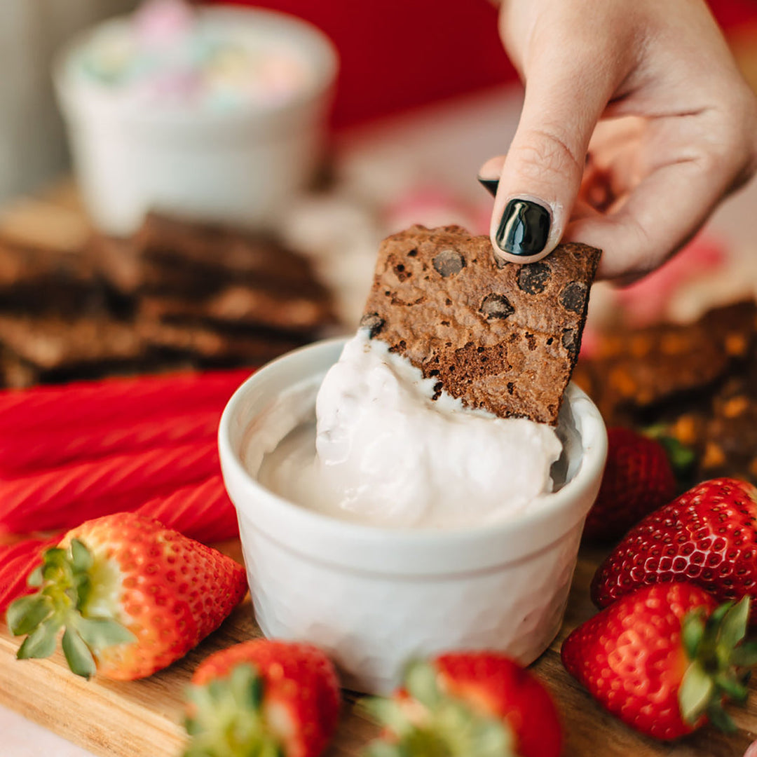 Person dipping a chocolate chip brownie brittle into a bowl of yogurt with strawberries around.