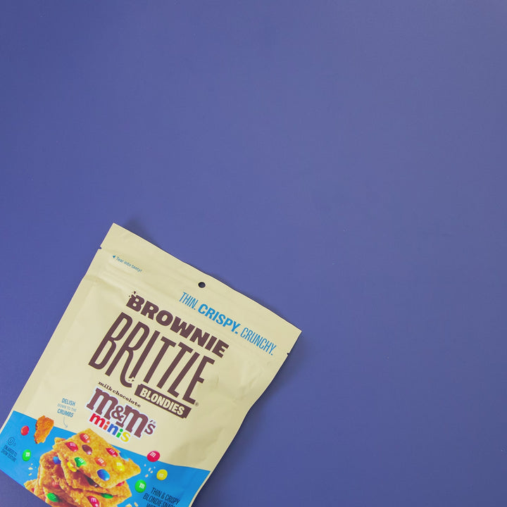 M&M'S® Minis Blondie Brownie Brittle Lifestyle Gif showing the pacge opening and brownie brittle spilling out over the table