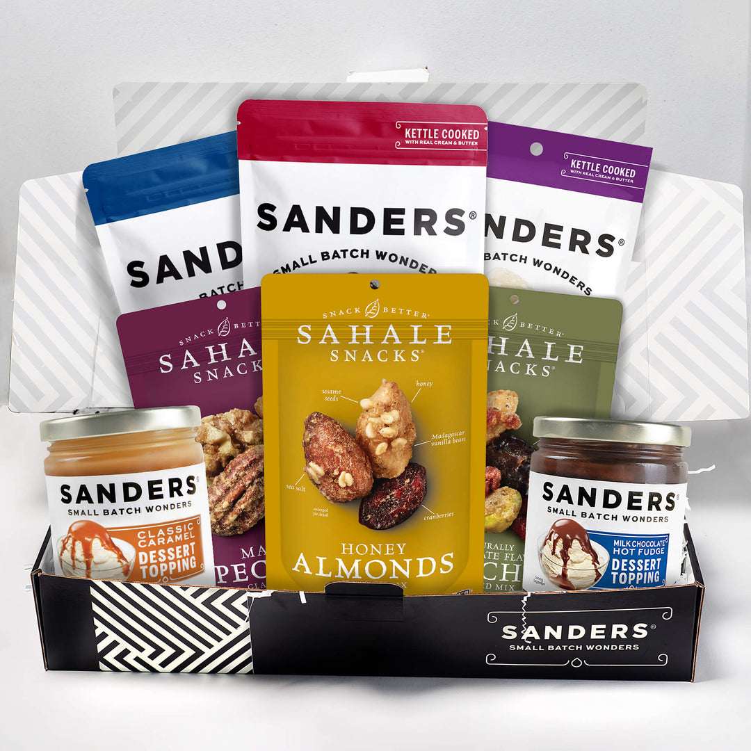 An assortment of snack products is displayed, including Sahale Snacks nut mixes in colorful packages and Sanders dessert toppings in glass jars, arranged in a gift box.